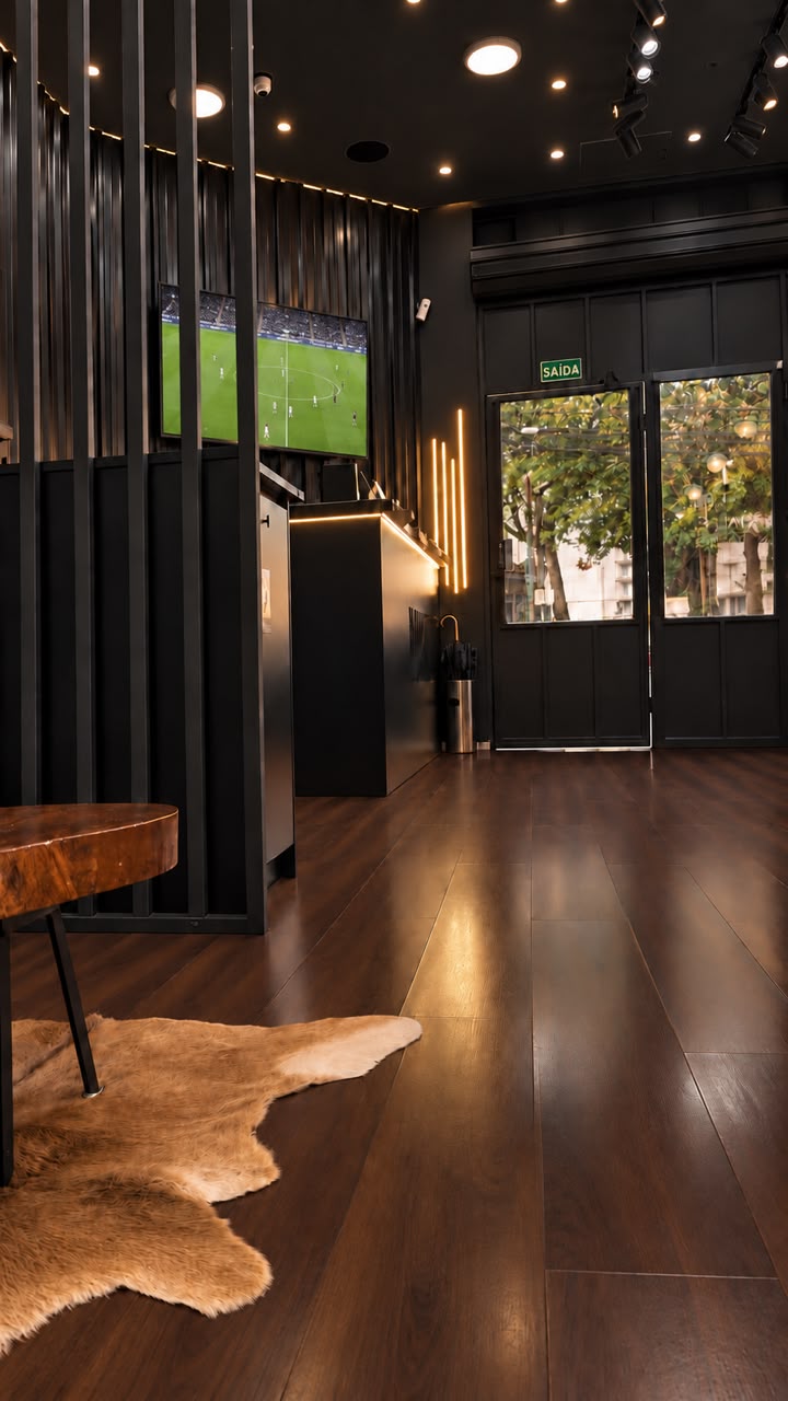 Recepção com piso de madeira, balcão iluminado e TV com futebol